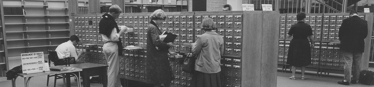 A black and white image of several people using a library card catalog.